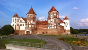 Dieses Archivfoto zeigt eine Außenansicht von Schloss Mir in Belarus im Sommer. Das Foto zeigt die malerische Festungsanlage mit vier Türmen hinter einem Blumenbeet und neben einem See.