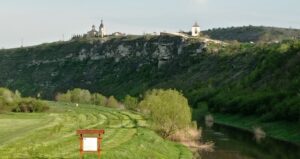 Ein Blick ins Grüne auf eine Weide neben einem Fluss im Vordergrund und auf eine Kirche, die sich am anderen Flussufer auf einer felsigen Erhebung befindet. Einen Eindruck von der landschaftlichen Schönheit Moldaus gewann die Gruppe auf der Fahrt aus dem Norden in die Hauptstadt Chișinău. Das Foto zeigt die Ausgrabungsstätte Orheiul Vechi (Altes Orhei) etwa 60 Kilometer nordöstlich der Haupstadt. Spuren von in die Felsen gegrabenen Festungen, Resten einer Moschee und christlichen Klöstern zeugen von der bewegten Vergangenheit der Gegend am Fluss Răut.