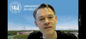Dieses Foto zeigt Dr. Roger Fornoff, Leiter des Lehrbereichs Deutsch als Fremdsprache an der Uni Köln, vor einem virtuellen Hintergrund der Universität, beim Vortrag im Projekt fokus plus: "Welche Werte dürfen wir erwarten?"