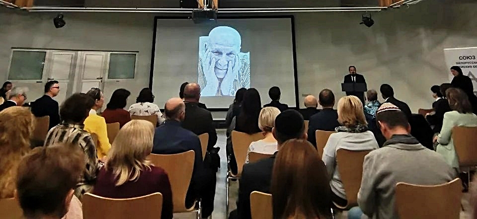 Ein Blick über die Köpfe des Publikums hinweg auf den Redner vor einer Leinwand, die ein Portrait von Leonid Lewin zeigt, auf dem er sehr warmherzig lächelt.
