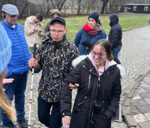 Ein blinder junger Mann wird von einer sehenden Frau über das unebene Kopfsteinpflaster der Gedenkstätte Stutthof geführt.