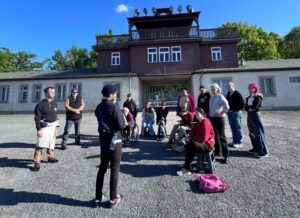 Auf dem Häftlingsgelände mit Blick auf das Eingangstor der Gedenkstätte steht die Gruppe im Halbkreis vor einer Person, die etwas erläutert. 