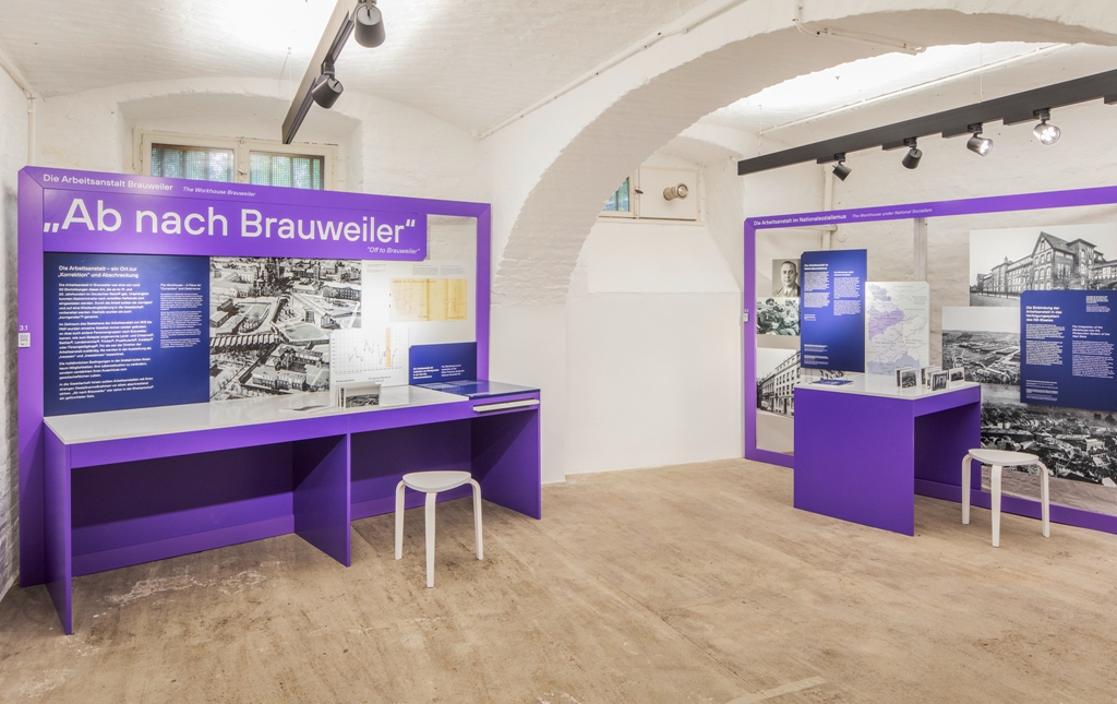 Das Foto zeigt einen Blick in einen Ausstellungsraum: Auf einer Tafel steht "Ab nach Brauweiler". Besucherinnen und Besucher können sich auf einen Hocker vor der Tafel setzen, um das große Foto genauer zu betrachten und um den langen Text zu lesen. 