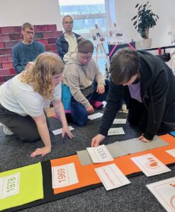 Mehrere Mitglieder knieen neben dem Zeitstrahl und sortieren die Text-Karten und Zeichnungen zu den verschiedenen Zeitabschnitten.