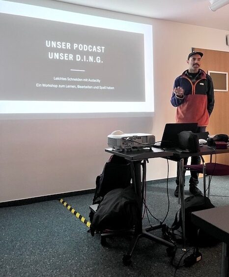 Rolando Gonzales steht neben einer Präsentation, die mit einem Beamer an die Wand geworfen wird und erklärt gerade etwas. 
