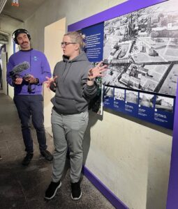 Eine Mitarbeiterin der Gedenkstätte Brauweiler steht für einer Ausstellungstafel mit einem großen Foto und mehreren kleineren und erläutert, was hier zu sehen ist. 