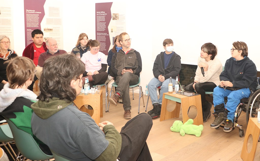 Das Foto zeigt neun Personen aus dem Podcast-Team im Halbkreis. Linn Bade vom Verein IN-Gesellschaft Berlin hat ein Mikrofon in der Hand und stellt gerade eine Frage.