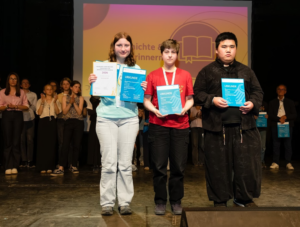 Marie, Mian und Jannik vom Podcast-Projekt "Das Erinnerungs-DING" stehen mit ihren Urkunden vorn auf der Bühne im Scheinwerferlicht. Sie haben die Urkunden stellvertretend für das ganze Team erhalten. Foto: Grit Hiersemann - demokratisch handeln